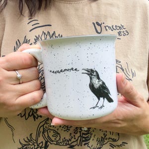 May include: White speckled ceramic mug with a black raven illustration and the word "nevermore". The mug has a black rim and a white handle. The mug is held by a person wearing a tan shirt.