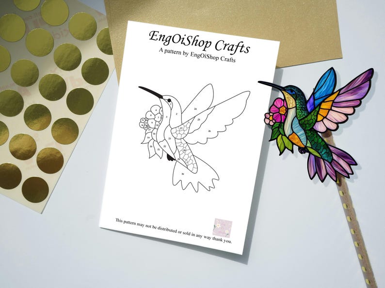 May include: A craft project featuring a hummingbird design. The image includes a black and white line drawing of a hummingbird with numbered sections, a colorful hummingbird decoration, and gold foil stickers. The text reads "EngOiShop Crafts".