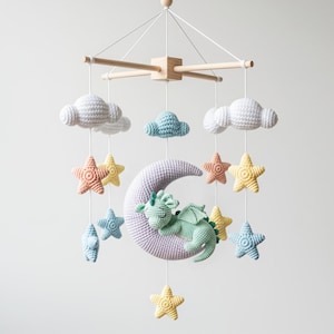 May include: A handmade baby mobile featuring a crocheted design. The mobile includes a light purple crescent moon with a sleeping green dragon, white clouds, and colorful stars in shades of peach, yellow, and blue. The wooden frame suspends the elements.
