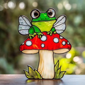 May include: A stained glass frog with wings sits on top of a red and white mushroom with a white stem. The mushroom is surrounded by green leaves.