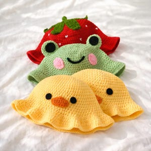 Puede incluir: Tres sombreros de pescador de ganchillo. El sombrero superior es rojo con un tallo verde y detalles blancos, que se asemeja a una fresa. El sombrero del medio es verde con rasgos de rana. Los sombreros inferiores son amarillos con rasgos de pato.