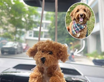May include: A plush, brown dog car charm hanging from a string, with a small, smiling dog portrait in the background. The charm has a fluffy texture and a small bow. The background shows a car interior and an outdoor scene.
