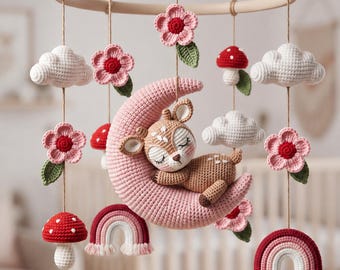 Könnte beinhalten: Ein Häkel-Mobile für Babys mit einem zentralen rehbraunen Reh, das auf einem rosa Halbmond schläft. Die hängenden Elemente umfassen weiße Wolken, rosa Blumen mit grünen Blättern, rot-weiße Pilze und kleine Regenbögen an einem Holzring.