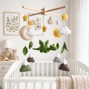May include: A handmade baby mobile with a wooden frame, featuring crocheted elements. The mobile includes a green dragon, yellow stars, a sun, clouds, mountains, leaves, and a moon. The mobile hangs above a white crib with a cream-colored blanket.