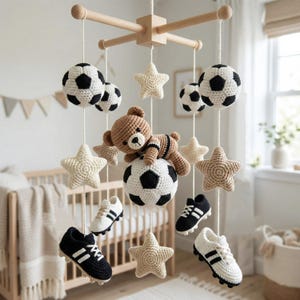 May include: A baby mobile with crochet soccer balls, stars, and miniature soccer cleats in black and white. A crochet teddy bear rests on a soccer ball. The mobile hangs from a wooden frame, a charming nursery decoration. The soccer cleats are approximately 10 cm long.