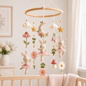 Op de afbeelding: Een hangende mobiel met een rond houten frame en gehaakte elementen, waaronder twee konijntjes in roze tutu's, een halve maan met bloemen, witte wolken, gele sterren, roze en witte bloemen, groene ranken en een rode paddenstoel met witte stippen.