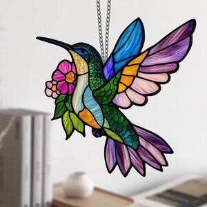 May include: A stained glass hummingbird with a long black beak, vibrant blue, green, yellow, and purple wings, and a pink flower. The bird is suspended by a silver chain, perfect for home decor.