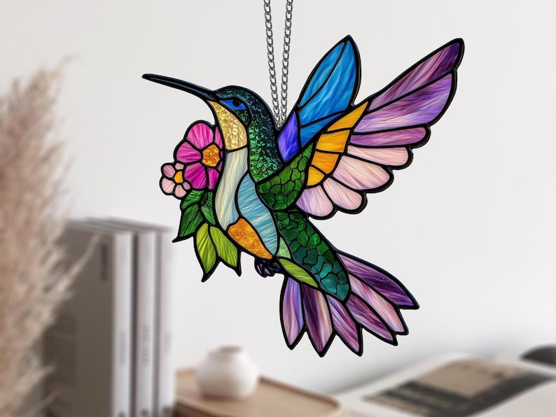 May include: A stained glass hummingbird with a long black beak, vibrant blue, green, yellow, and purple wings, and a pink flower. The bird is suspended by a silver chain, perfect for home decor.