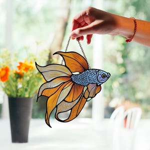 May include: A stained glass window hanging with a blue and orange fish design. The fish has a long, flowing tail and fins.