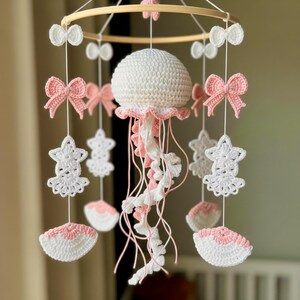 May include: A handmade baby mobile featuring a crocheted jellyfish with white and pink details. The mobile includes crocheted bows, stars, and shell shapes, all suspended from a wooden hoop. The color palette is soft and pastel.