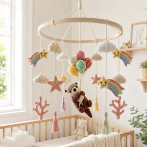 May include: A handmade baby mobile featuring crocheted elements. The mobile includes a wooden ring, clouds, stars, rainbows, an otter, coral, and balloons in pastel colors. The mobile hangs above a wooden crib.