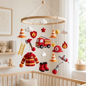 May include: A nursery mobile featuring crocheted fire-themed elements suspended from a circular wooden frame. The hanging items include a red fire truck, a yellow ladder, orange traffic cones, a firefighter jacket, boots, an axe, and white clouds.