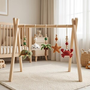 May include: A wooden baby play gym with hanging crochet toys. The toys include a turtle, cloud, palm tree, starfish, and crab. The toys are in shades of green, orange, and red, with wooden beads. The play gym is made of light-colored wood.