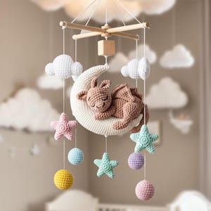 May include: A handmade baby mobile featuring a crocheted dragon sleeping on a crescent moon. The mobile includes white clouds, pastel stars, and colorful spheres. The wooden frame suspends the soft, textured elements, creating a whimsical nursery decoration.