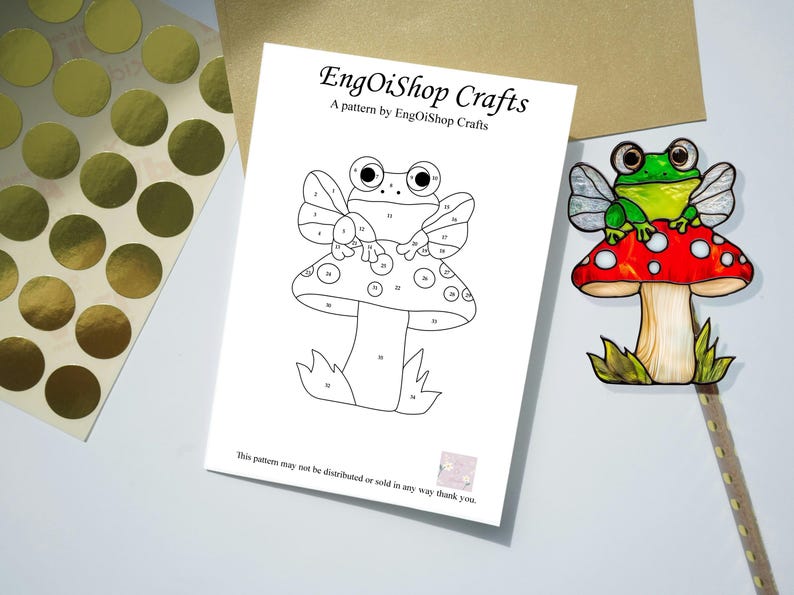 May include: A black and white line art pattern of a frog sitting on a mushroom with wings. The pattern is numbered for a paint-by-number craft project. The text "EngOiShop Crafts" and "A pattern by EngOiShop Crafts" is at the top of the page. The text "This pattern may not be distributed or sold in any way thank you." is at the bottom of the page.