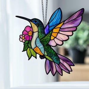 May include: A stained glass hummingbird with a long black beak, holding a pink flower. The bird's wings are blue, yellow, and purple. The body is green and gold, with a white chest. It hangs from a silver chain.