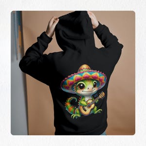 Puede incluir: Sudadera negra con un lagarto verde que lleva un sombrero de colores y toca la guitarra. El lagarto tiene ojos grandes y una sonrisa amigable.