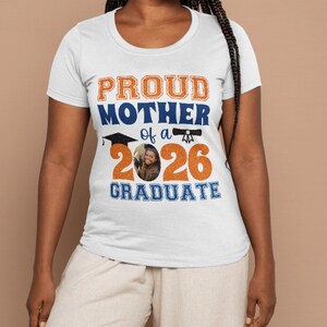 以下が含まれることがあります： オレンジと青で「PROUD MOTHER of a 2026 GRADUATE」と書かれた白いTシャツ。卒業キャップと2026年の数字の中に人の写真がプリントされています。