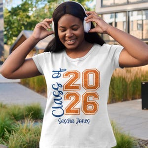 以下が含まれることがあります： 白のTシャツ。青い筆記体とオレンジ色のブロック文字で「Class of 2026」と書かれています。名前の「Sascha Jones」は下に印刷されています。卒業をテーマにしたデザインです。