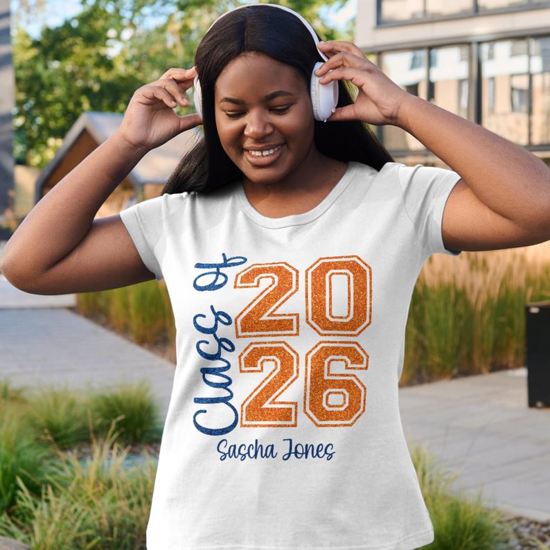 以下が含まれることがあります： 白のTシャツ。青い筆記体とオレンジ色のブロック文字で「Class of 2026」と書かれています。名前の「Sascha Jones」は下に印刷されています。卒業をテーマにしたデザインです。