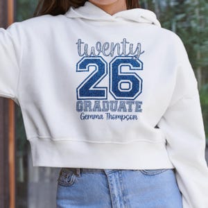以下が含まれることがあります： 白いフード付きスウェットシャツ。前面に青いグリッター文字で「twenty 26 GRADUATE Gemma Thompson」と書かれています。スウェットシャツはクロップドスタイルで、青いジーンズと組み合わせています。デザインはスウェットシャツの中央に配置されています。