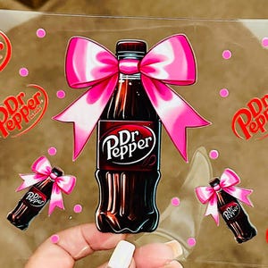 May include: A sheet of clear vinyl with multiple Dr Pepper logos and images of Dr Pepper bottles with pink bows. The bottles are brown and the bows are pink with white polka dots.