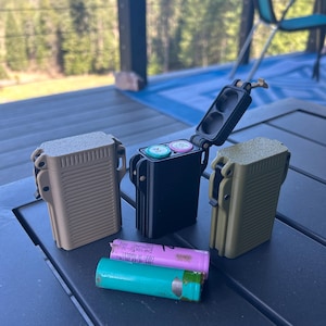 May include: Three small, rectangular, hinged cases in beige, black, and green, with one case open revealing two small, round, colored battery compartments. Two cylindrical batteries, one pink and one turquoise, are lying on a dark gray surface.