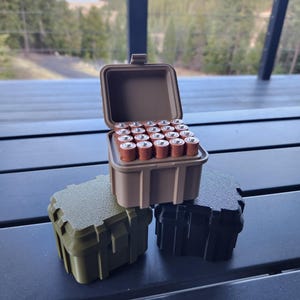 May include: Three small, stackable, plastic storage containers. The top container is tan and open, revealing a compartment filled with brown and silver AA batteries. The bottom two containers are green and black.