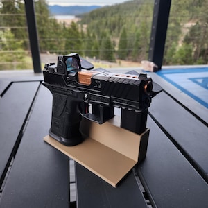 May include: A black handgun with a red dot sight and a tan colored base. The gun is on a wooden table with a view of a forest and a lake in the background.