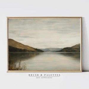 May include: A landscape painting in a gold frame depicting a calm lake surrounded by rolling hills under a soft, overcast sky. The artwork features muted tones of brown, green, and grey, with the text BRUSH & PALETTES art collective below.