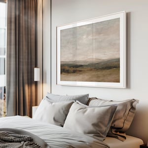 May include: A framed landscape painting with a neutral color palette hangs above a bed with gray pillows and bedding. The artwork depicts a field, hills, and a cloudy sky. The frame is a light metallic color.