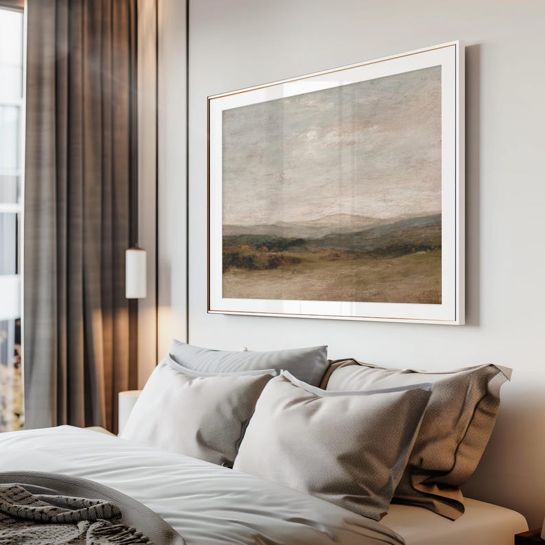 May include: A framed landscape painting with a neutral color palette hangs above a bed with gray pillows and bedding. The artwork depicts a field, hills, and a cloudy sky. The frame is a light metallic color.