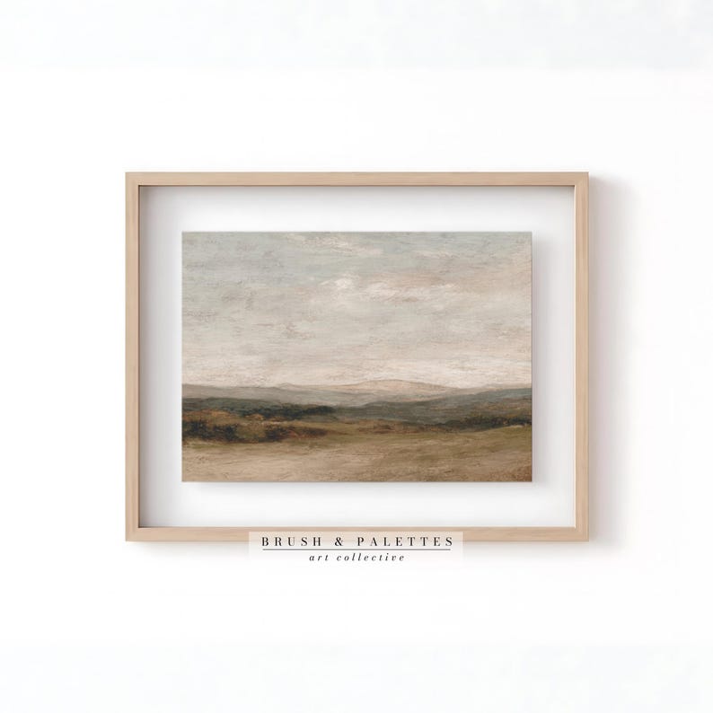 May include: Framed landscape art print featuring a muted color palette of beige, brown, and gray. The artwork depicts a serene countryside scene with rolling hills and a cloudy sky. The frame is a light wood color. The text "BRUSH & PALETTES art collective" is at the bottom.