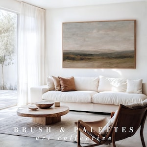 May include: A living room with a large landscape painting in a gold frame above a white sofa with brown throw pillows. A wooden coffee table and a leather chair are in front of the sofa. The text "BRUSH & PALETTES art collective" is at the bottom.
