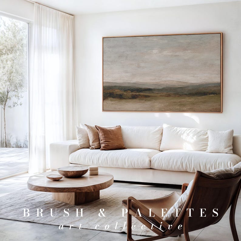 May include: A living room with a large landscape painting in a gold frame above a white sofa with brown throw pillows. A wooden coffee table and a leather chair are in front of the sofa. The text "BRUSH & PALETTES art collective" is at the bottom.