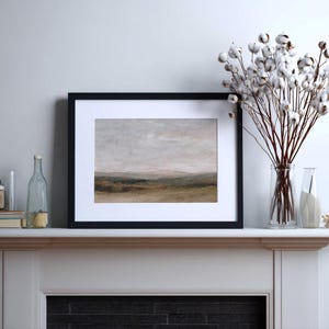 May include: A framed landscape art print with a black frame and white matting. The artwork depicts a muted, neutral-toned landscape with rolling hills and a cloudy sky. A vase of cotton stems sits to the right.