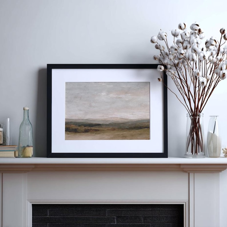 May include: A framed landscape art print with a black frame and white matting. The artwork depicts a muted, neutral-toned landscape with rolling hills and a cloudy sky. A vase of cotton stems sits to the right.