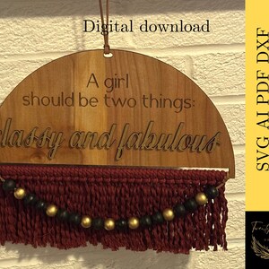 May include: A wooden half-circle sign with the text "A girl should be two things: classy and fabulous" in a cursive font. The sign is hanging from a string and has a burgundy tassel with gold and black beads.