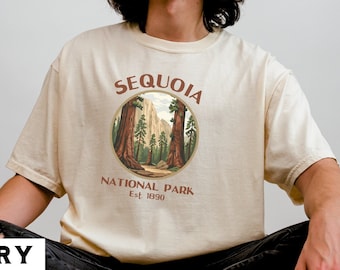 May include: A person wearing a cream colored t-shirt with a brown circle graphic that says "Sequoia National Park Est. 1890". The graphic features a depiction of a forest with tall trees.
