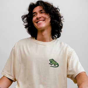 May include: Ivory-colored XL t-shirt with a green iguana graphic. The model is 5'10". The shirt is a neutral color and has a relaxed fit. The iguana graphic is on the left chest. The shirt is made of a soft material.