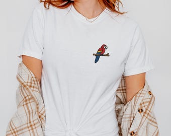 May include: White t-shirt with a small embroidered parrot design on the chest. The parrot is red, blue, and yellow, perched on a brown branch. The shirt is paired with blue jeans and a plaid shirt. The text "WHITE" and "MODEL: 5'4", SIZE: S" are visible.