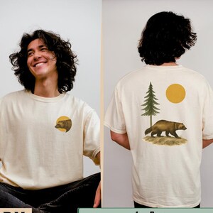 May include: Ivory-coloured t-shirt with a round neck. The front has a circular graphic of a bear's head. The back displays a wolverine, a tree, and a sun graphic. The text "IVORY" and "MODEL: 5'10", SIZE: XL" are also visible.