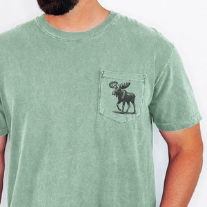 Puede incluir: Una camiseta verde salvia con un gráfico de alce en el bolsillo. El modelo mide 1,83 m de altura y lleva una talla L.