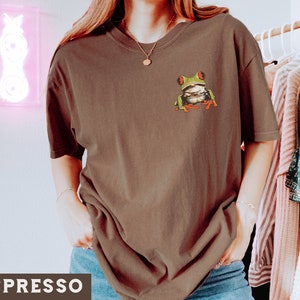 May include: A brown t-shirt with a green tree frog graphic on the front. The frog has red eyes and is sitting on a branch.