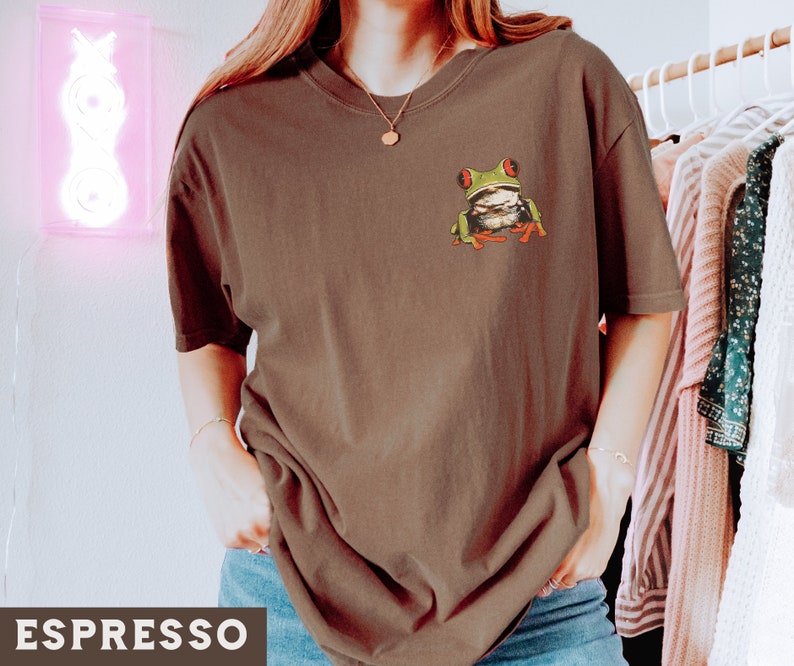 May include: A brown t-shirt with a green tree frog graphic on the front. The frog has red eyes and is sitting on a branch.