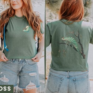 May include: A green t-shirt with a chameleon graphic on the back and a small graphic on the front. The t-shirt is being worn by a person with long red hair. The person is wearing blue denim shorts.