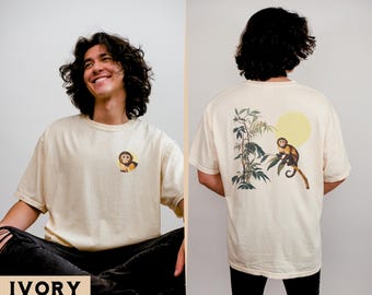 Op de afbeelding: Een persoon draagt een licht ivoorwit T-shirt met een klein apenmotief aan de voorkant en een groter apenmotief aan de achterkant. Het motief aan de achterkant toont een aap die op een tak zit met bladeren en een gele zon.