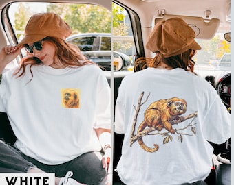 Puede incluir: Camiseta blanca con una pequeña imagen de un mono en el lado izquierdo y una imagen más grande de un mono en el lado derecho. El mono es marrón y tiene una cola larga.