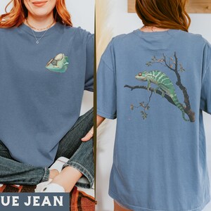 May include: A blue jean t-shirt with a chameleon graphic on the front and a larger chameleon graphic on the back. The chameleon on the back is on a branch with leaves and berries.