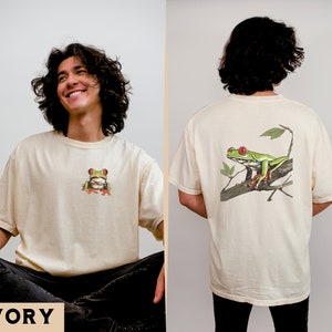 May include: A cream colored t-shirt with a green tree frog on the front and a larger green tree frog on the back. The text "IVORY" is at the bottom of the image.